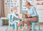 Young smiling caucasian child psychologist sitting with an adorable little girl in a clinic and using a clipboard for a consult. Cute girl talking to a mental health professional. Dyslexia and anxiety