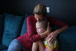 Woman and sad little daughter are sitting on couch and hugging